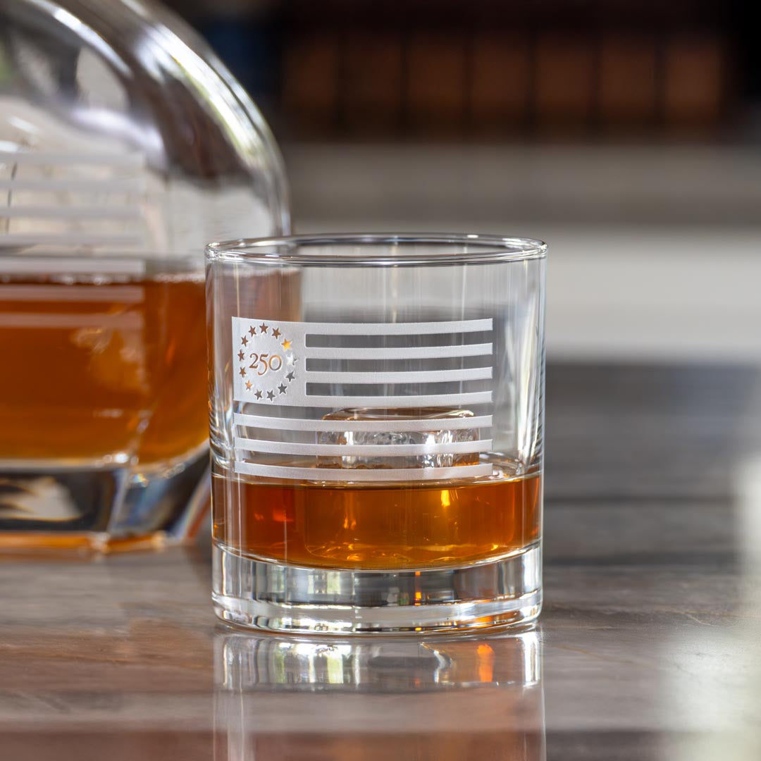 Clear whiskey glass with etched Betsy Ross flag design containing amber liquid on reflective surface