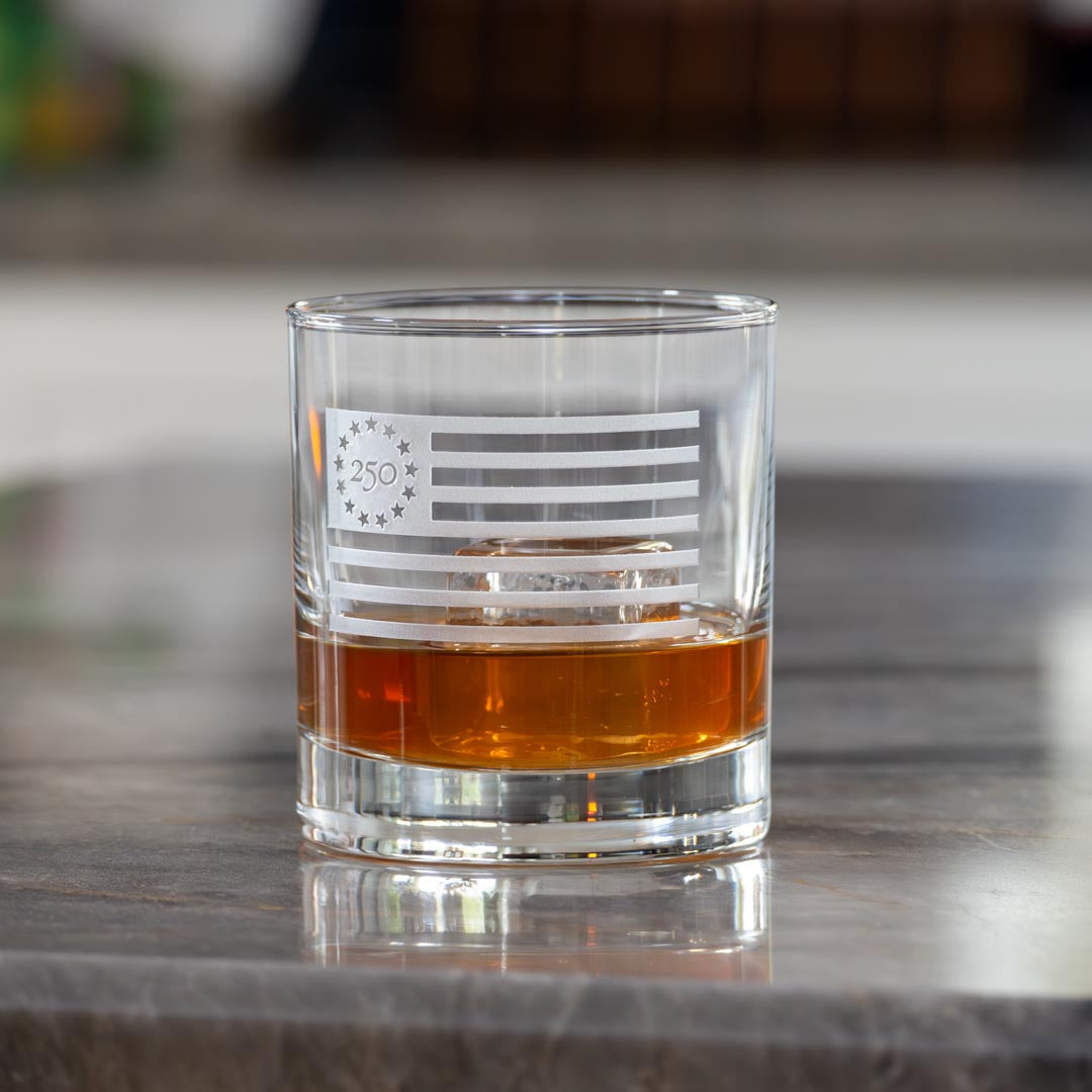 Clear whiskey glass with Betsy Ross flag design containing amber liquid on marble surface