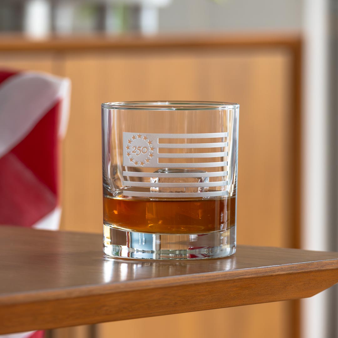 Clear whiskey glass with Betsy Ross flag design containing amber liquid on wooden surface
