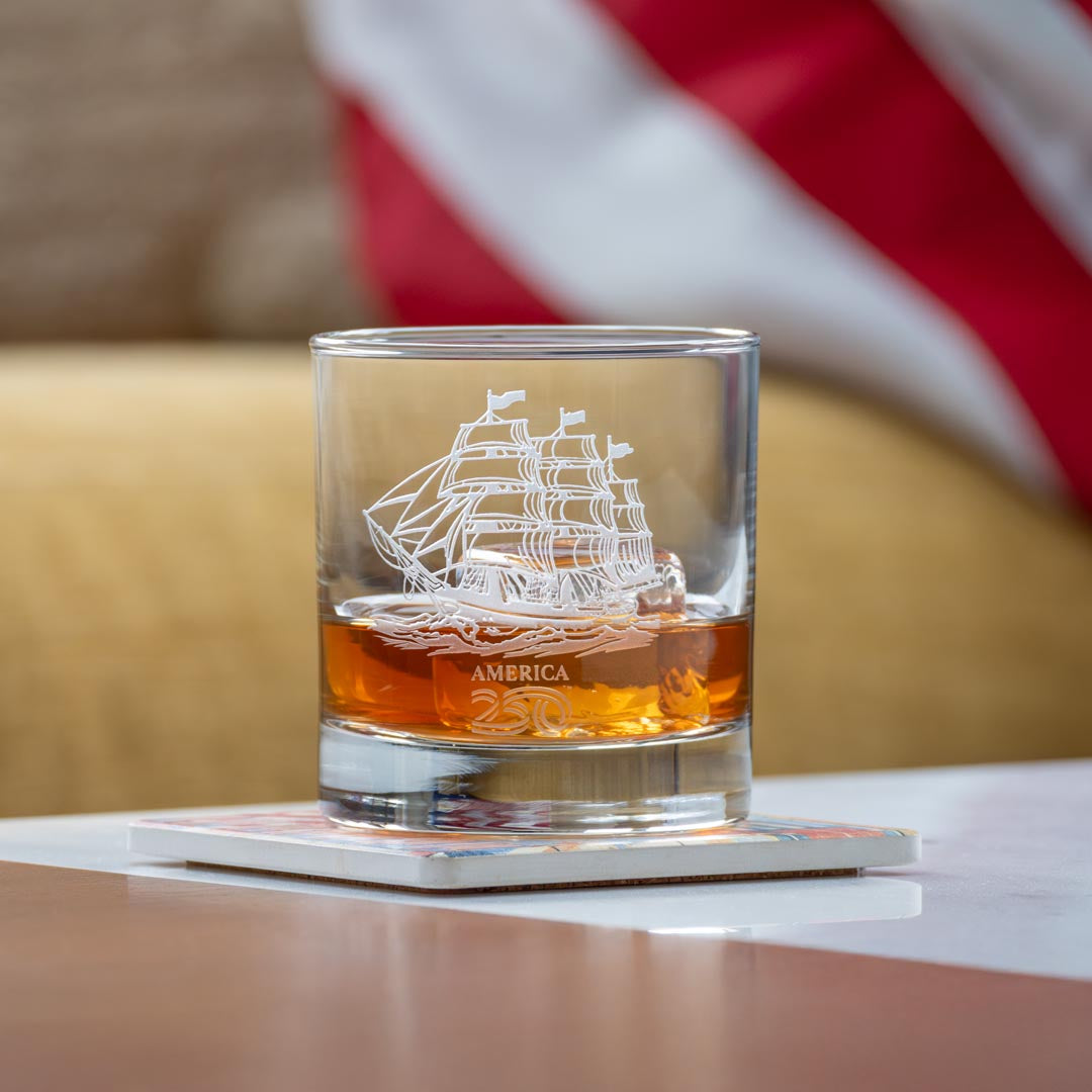 Clear whiskey glass with etched tall ship design containing amber whiskey on reflective surface