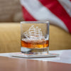 Clear whiskey glass with etched tall ship design containing amber whiskey on reflective surface