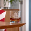 Clear whiskey glass with etched tall ship design containing amber whiskey on wooden surface