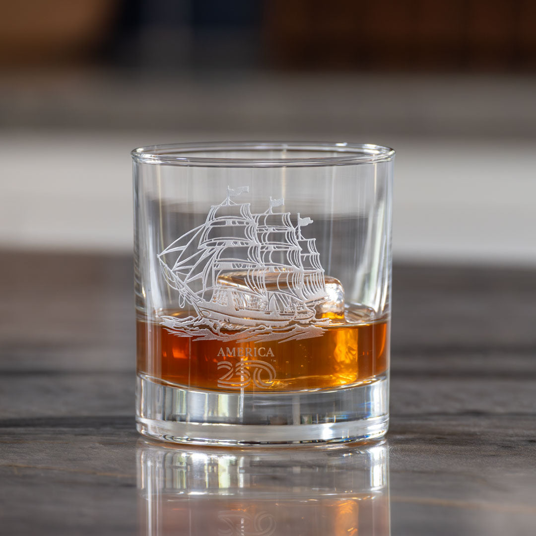 Clear glass whiskey tumbler with etched tall ship design containing amber colored whiskey