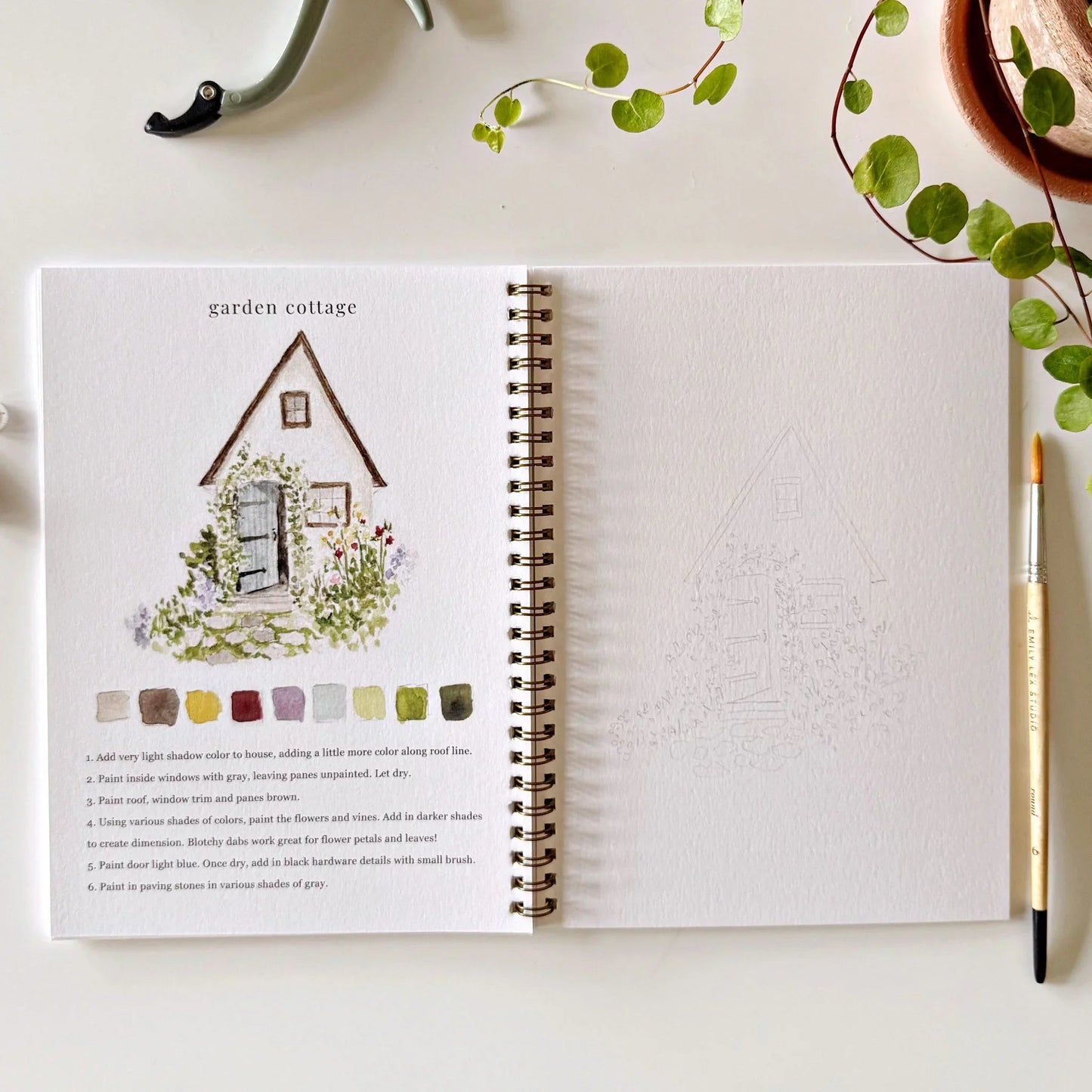 Open watercolor workbook showing garden cottage tutorial with paint swatches and blank practice page