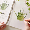 Hands painting watercolor watering can in green spiral-bound garden workbook with paint swatches
