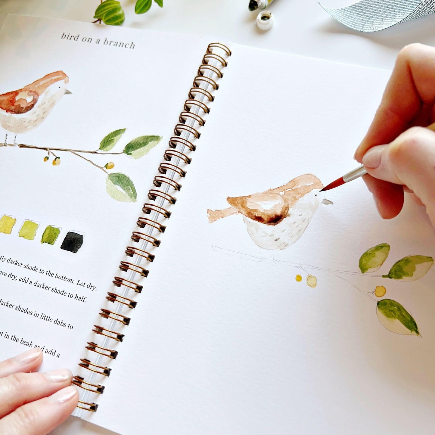 Hand painting watercolor bird in open spiral-bound workbook with nature illustrations and color swatches