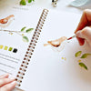 Hand painting watercolor bird in open spiral-bound workbook with nature illustrations and color swatches