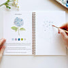 Open watercolor workbook showing hydrangea painting tutorial with hands holding paintbrush and color swatches