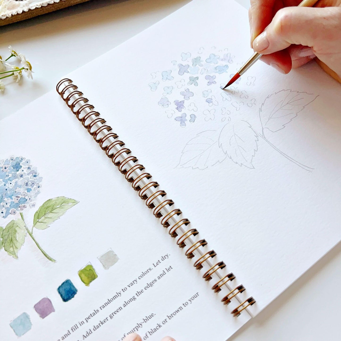 Hand painting watercolor flowers in spiral-bound workbook with botanical illustrations and color swatches