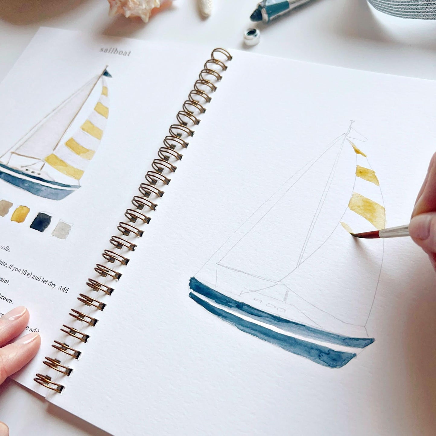 Open watercolor workbook showing sailboat painting tutorial with color swatches and brush in hand
